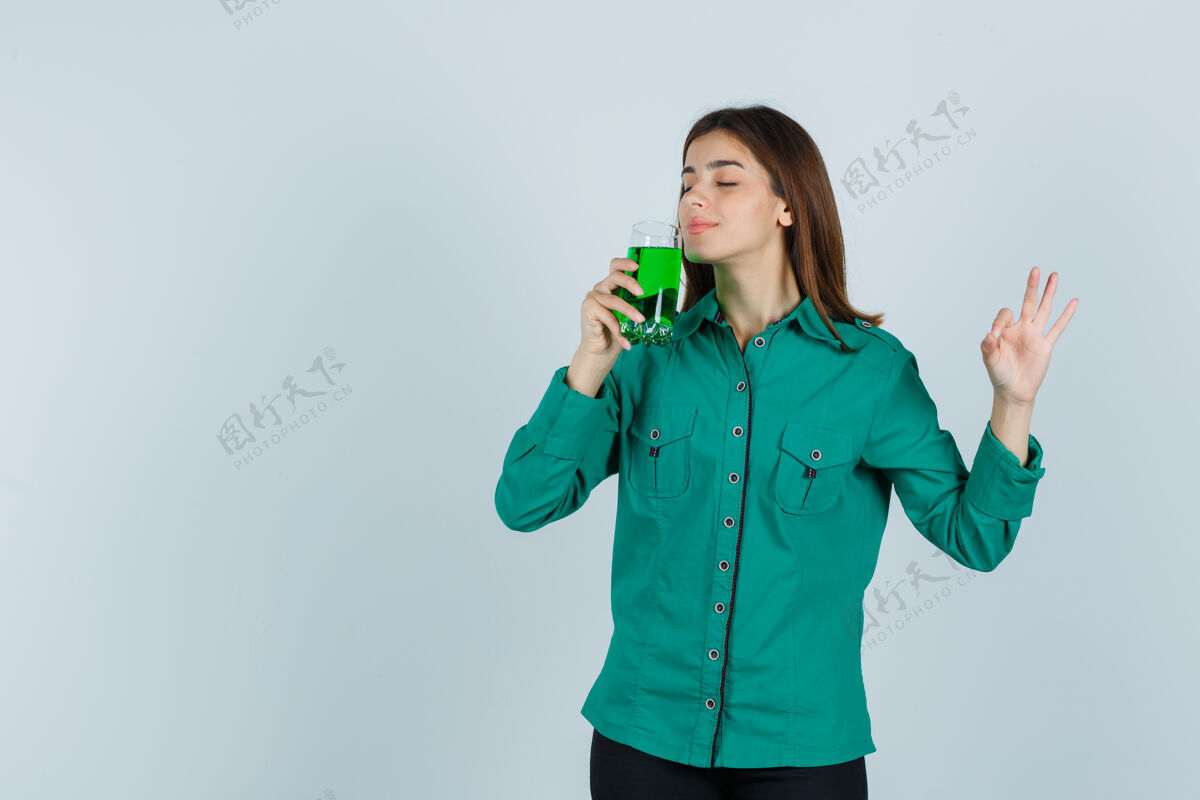 衬衫年轻女孩闻到一杯绿色液体的味道 穿着绿色上衣 黑色裤子 看上去很乐观 正面照女士时尚女孩