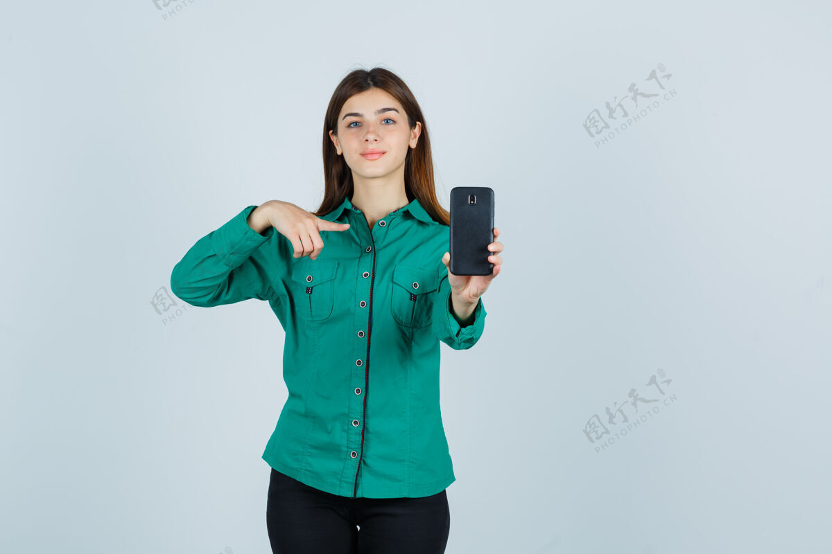 人穿着绿色上衣 黑色裤子的年轻女孩 一手拿着手机 用食指指着手机 神情欢快 正对着前方时尚休闲女性