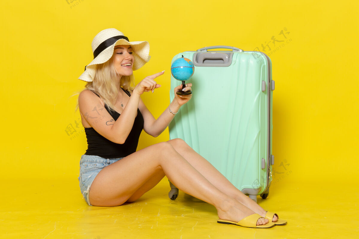 海正面图度假中的年轻女性坐在她绿色的袋子里拿着地球仪在黄色的墙上女性旅行旅行海色太阳健身运动吹风机