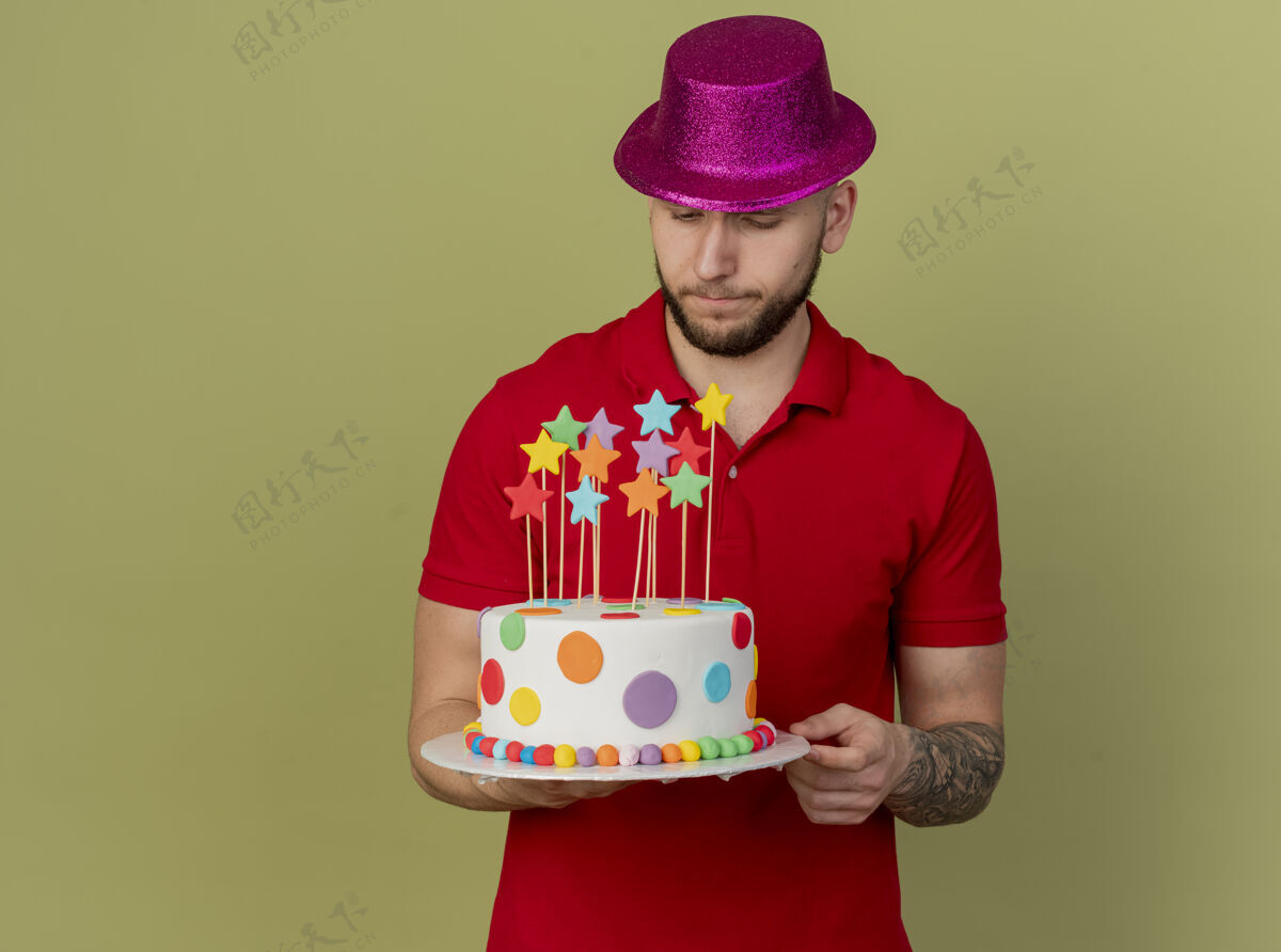 年轻人体贴的年轻帅哥斯拉夫党家伙戴着党的帽子举行 看着生日蛋糕孤立的橄榄绿背景与复制空间斯拉夫人空间小伙子