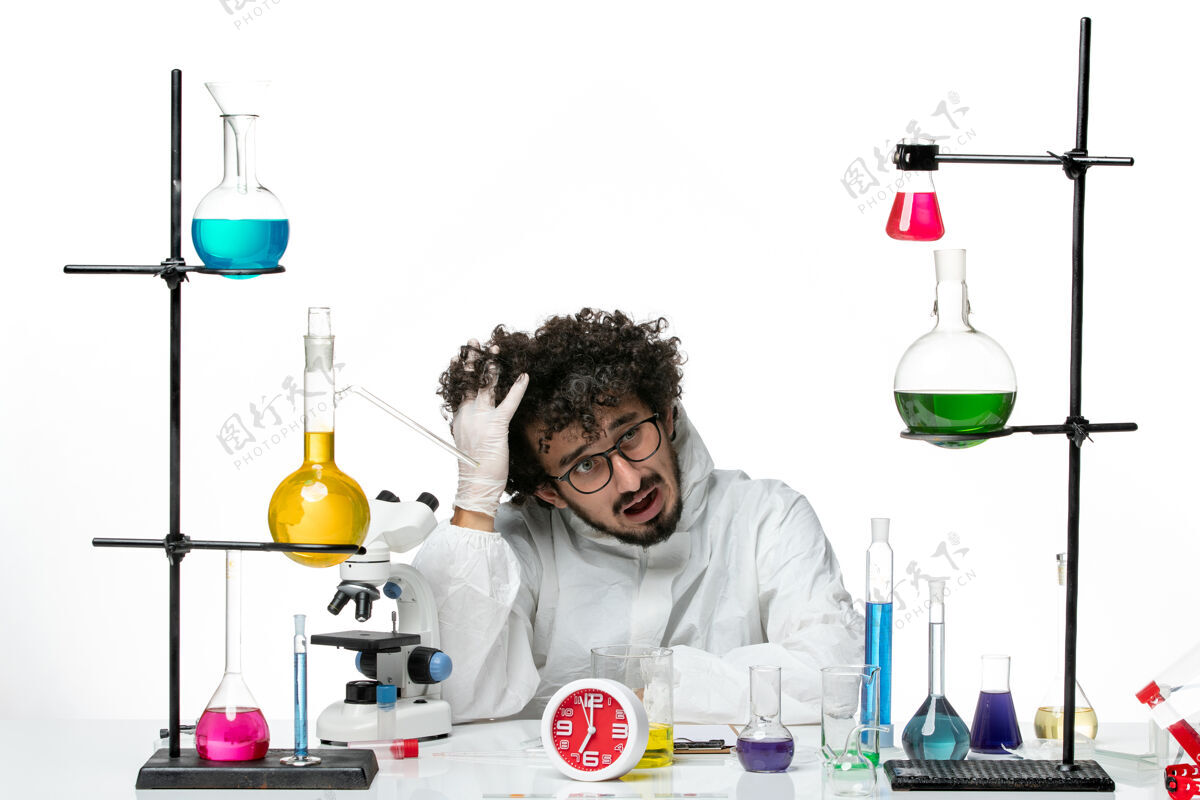 溶液正面图穿着特殊套装的年轻男性科学家坐在白墙上 拿着解决方案科学化学烧杯