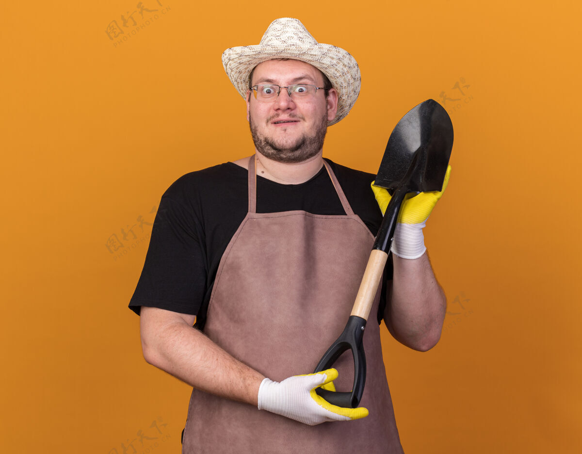 帽子惊讶的年轻男园丁戴着园艺帽和手套 手持铁锹隔离在橙色的墙上黑桃手套穿