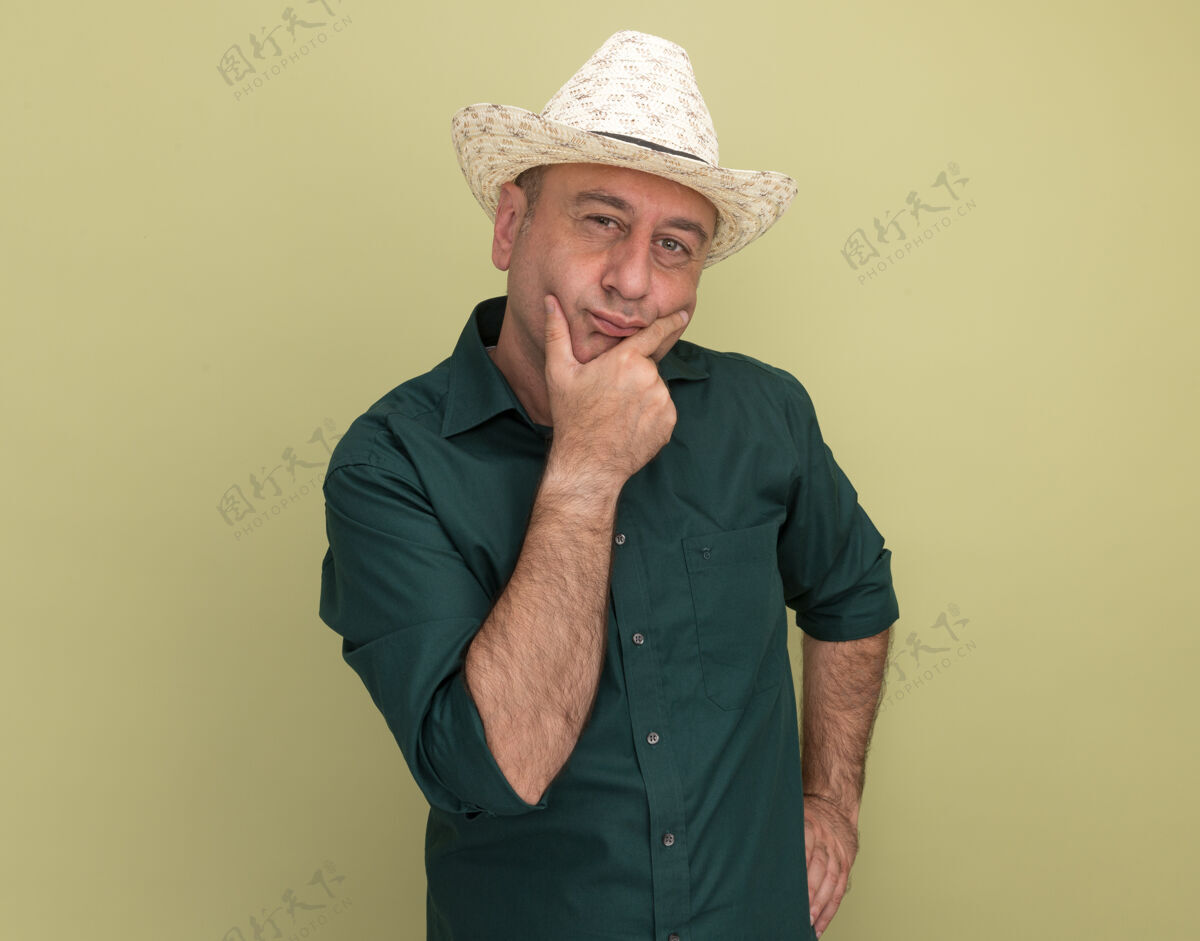 抓住想着中年男子穿着绿色t恤 戴着帽子 抓着下巴把手放在腰上 隔离在橄榄绿的墙上男人T恤帽子