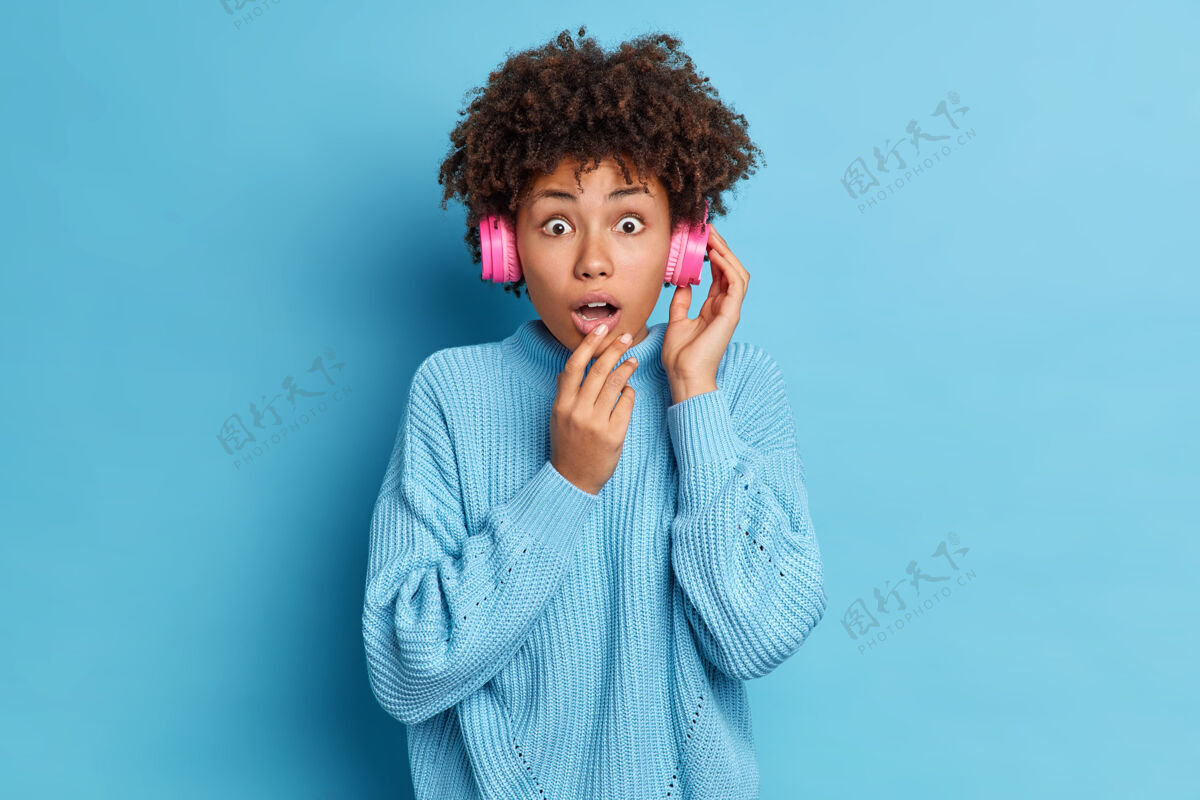 时髦说不出话的黑皮肤美国黑人年轻女子给人留下深刻印象的面部表情戴着立体声耳机穿着超大号针织毛衣张嘴耳机声音种族