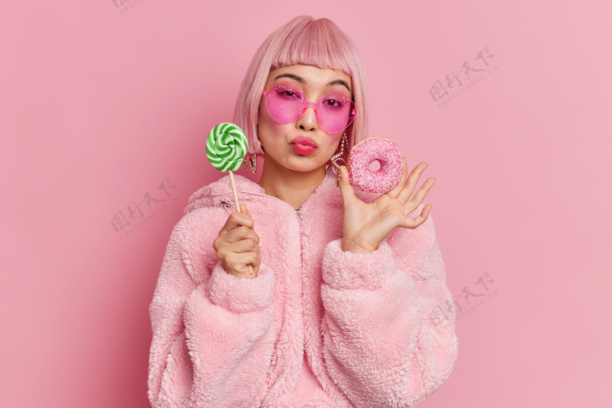 休闲浪漫时尚的粉红头发迷人的女模特爱吃甜食拿着棒棒糖和釉面甜甜圈穿着暖和的皮草外套时髦的心形太阳镜皮草浪漫满意