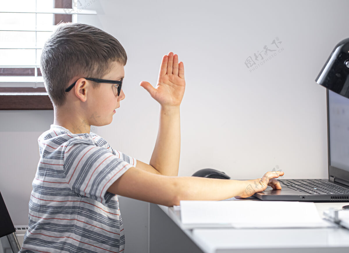 学生一个戴眼镜的小学生坐在桌旁 拿着笔记本电脑 远程学习 在网上课上举手教育学生桌子