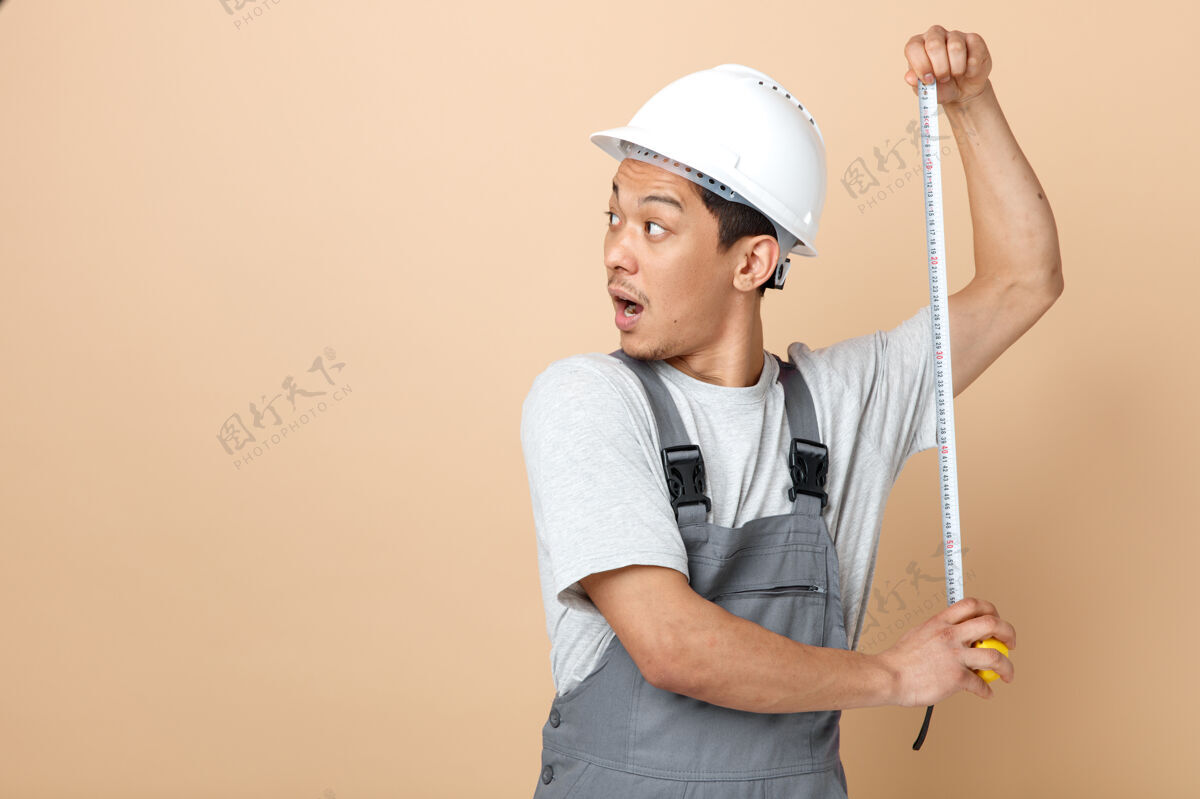 头盔印象深刻的年轻建筑工人戴着安全帽和制服拿着卷尺看着旁边持有建筑壁板