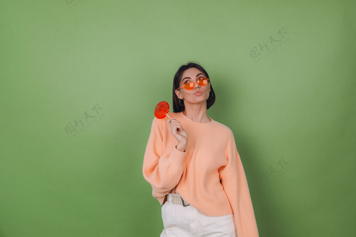 女性年轻时尚的女士穿着休闲的桃色毛衣 戴着橘色眼镜 隔离在绿色橄榄色的墙上 用橘色棒棒糖若有所思地看着一边思考着复制空间化妆品焦糖青年