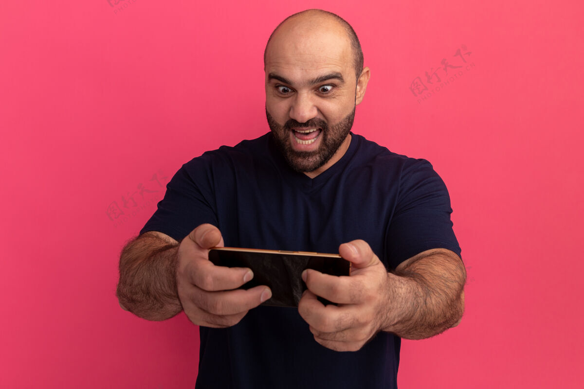男人留着胡子的男人穿着海军蓝t恤 用智能手机玩游戏快乐而情绪激动地站在粉色的墙上游戏情绪胡子