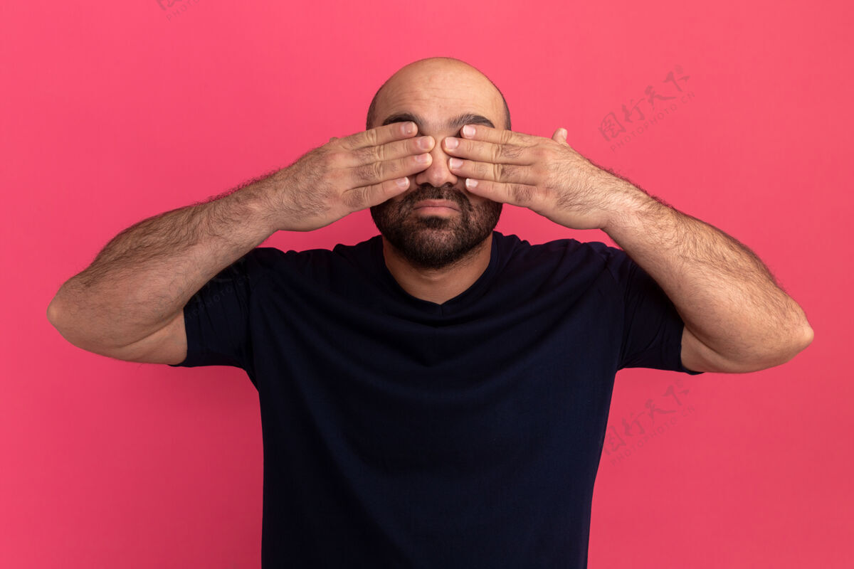 站起来留着胡子的男人穿着海军蓝t恤 双手站在粉色的墙上遮住眼睛胡子掩护伙计
