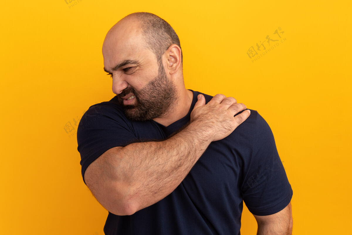 不舒服一个留着胡子的男人穿着海军蓝t恤 看起来不舒服 摸着肩膀 站在橘色的墙上感到疼痛胡须感觉男人