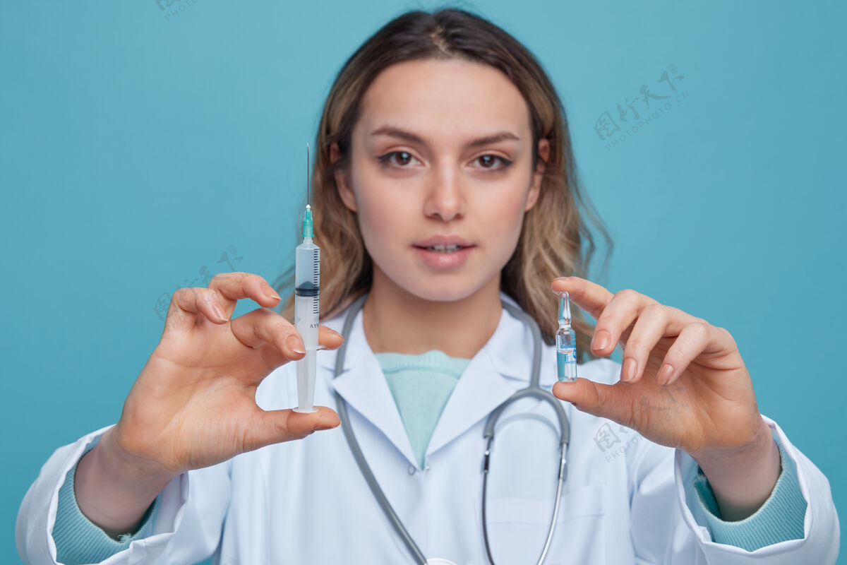 蓝色特写镜头自信的年轻女医生穿着医用长袍 脖子上戴着听诊器 向镜头伸出注射器和安瓿特写穿着长袍