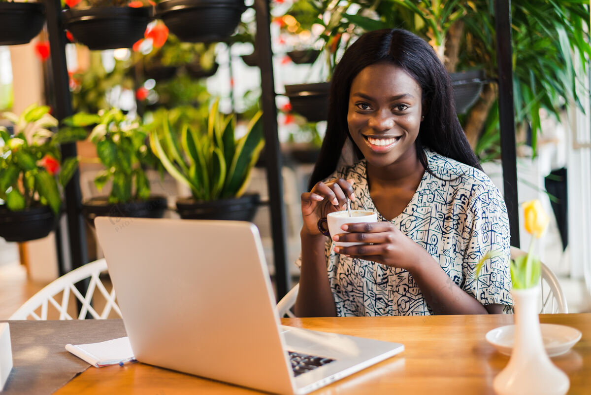 人一位非洲裔美国妇女拿着笔记本电脑和咖啡在咖啡馆放松的画像表情持有香气