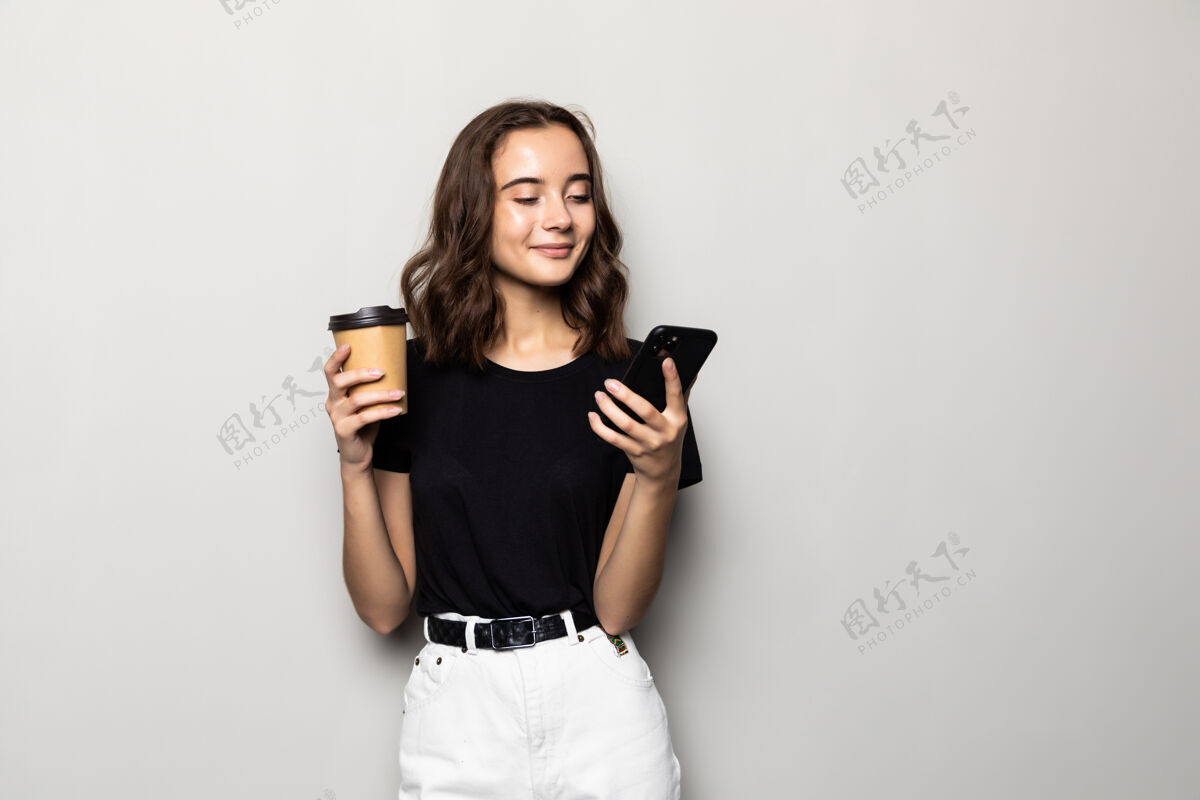 人一位穿着正式服装的成功女性站在灰色的墙上 手里拿着智能手机和外卖咖啡手机咖啡请
