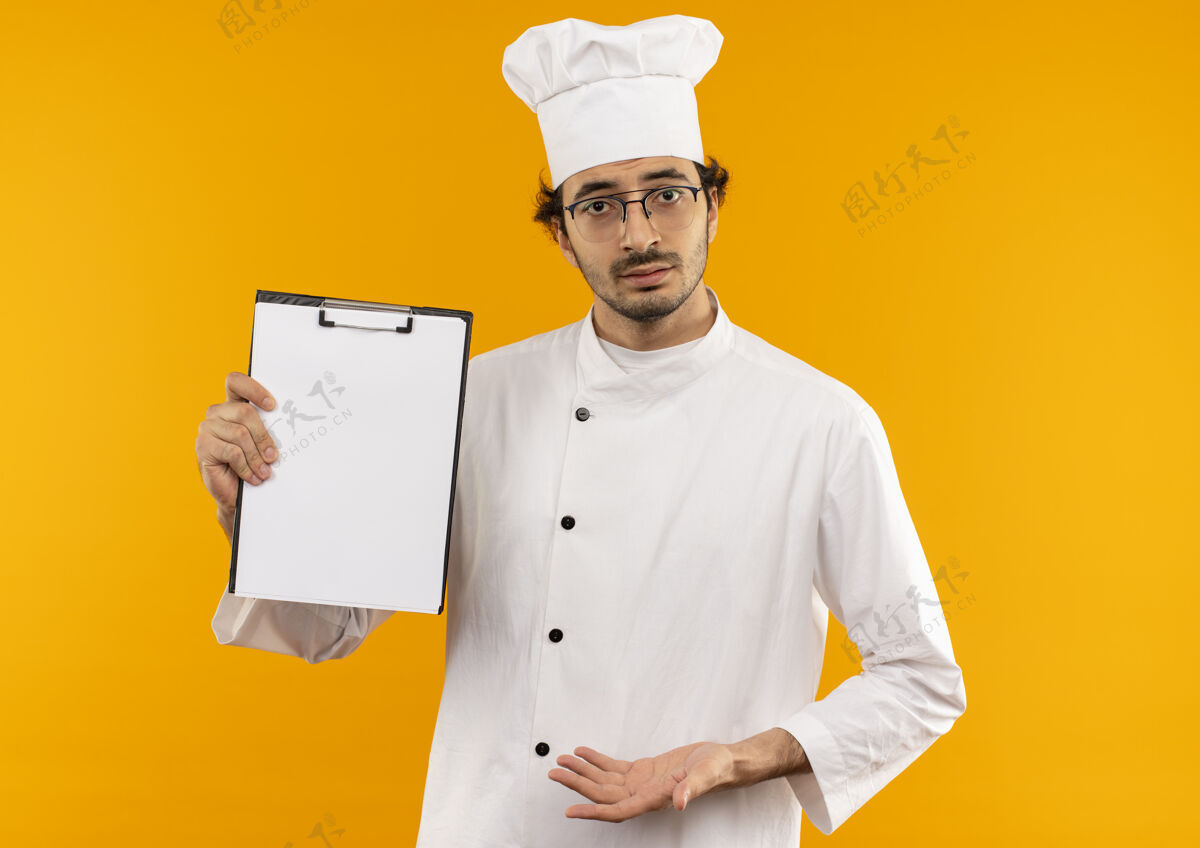 烹饪困惑的年轻男厨师穿着厨师制服 戴着眼镜拿着剪贴板混乱剪贴板黄色