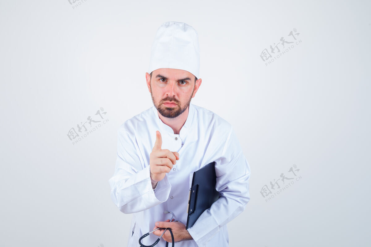 肖像身穿白色制服的年轻男医生拿着剪贴板 听诊器 用手指警告 表情严肃 正前方视图人男性制服