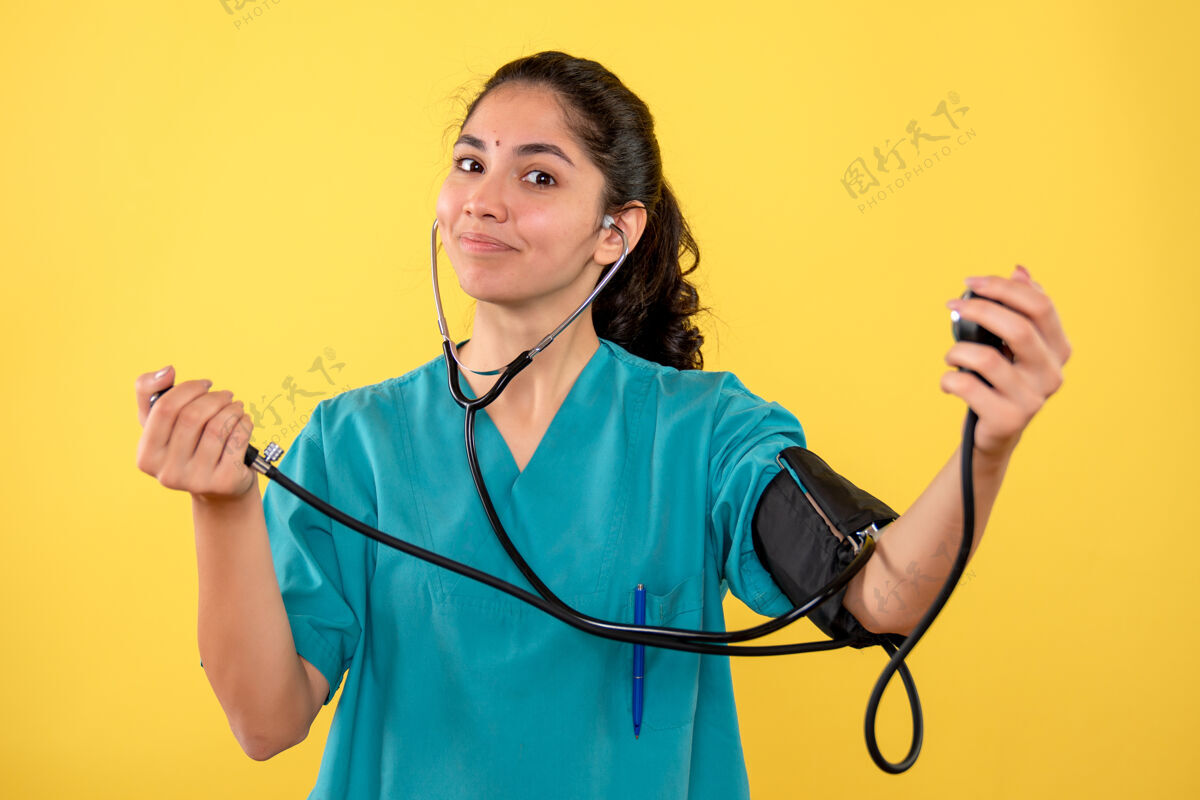 手持正面图身着制服的快乐女医生手持血压计站在黄色背景上背景护士医学