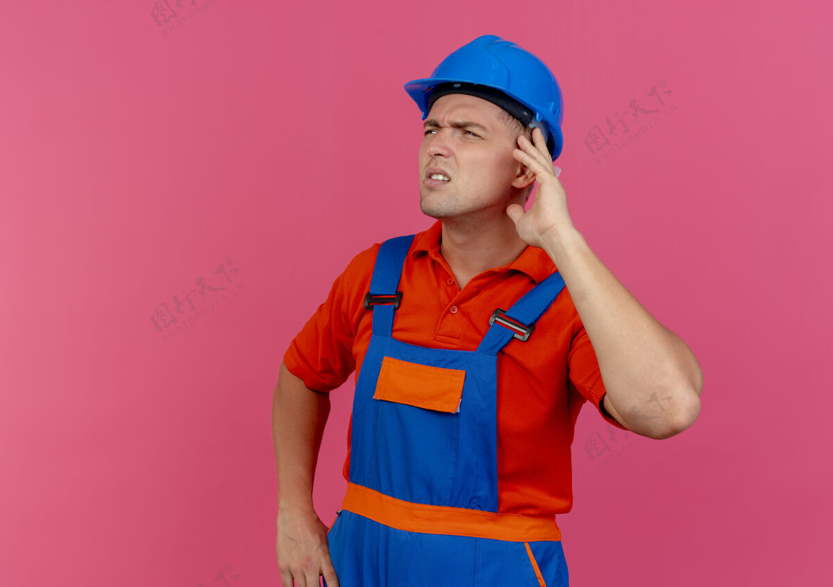 思想看着一边思考的年轻男建筑工人穿着制服 戴着安全帽 手放在耳朵上手粉色耳朵