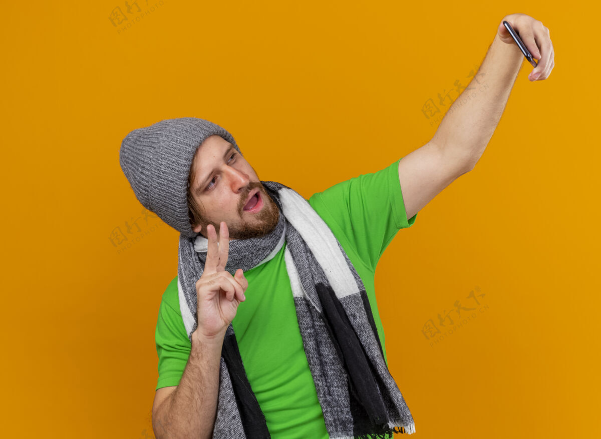 橙色自信的年轻英俊的斯拉夫坏人戴着冬天的帽子和围巾自拍做和平标志隔离在橙色的墙上围巾疾病穿