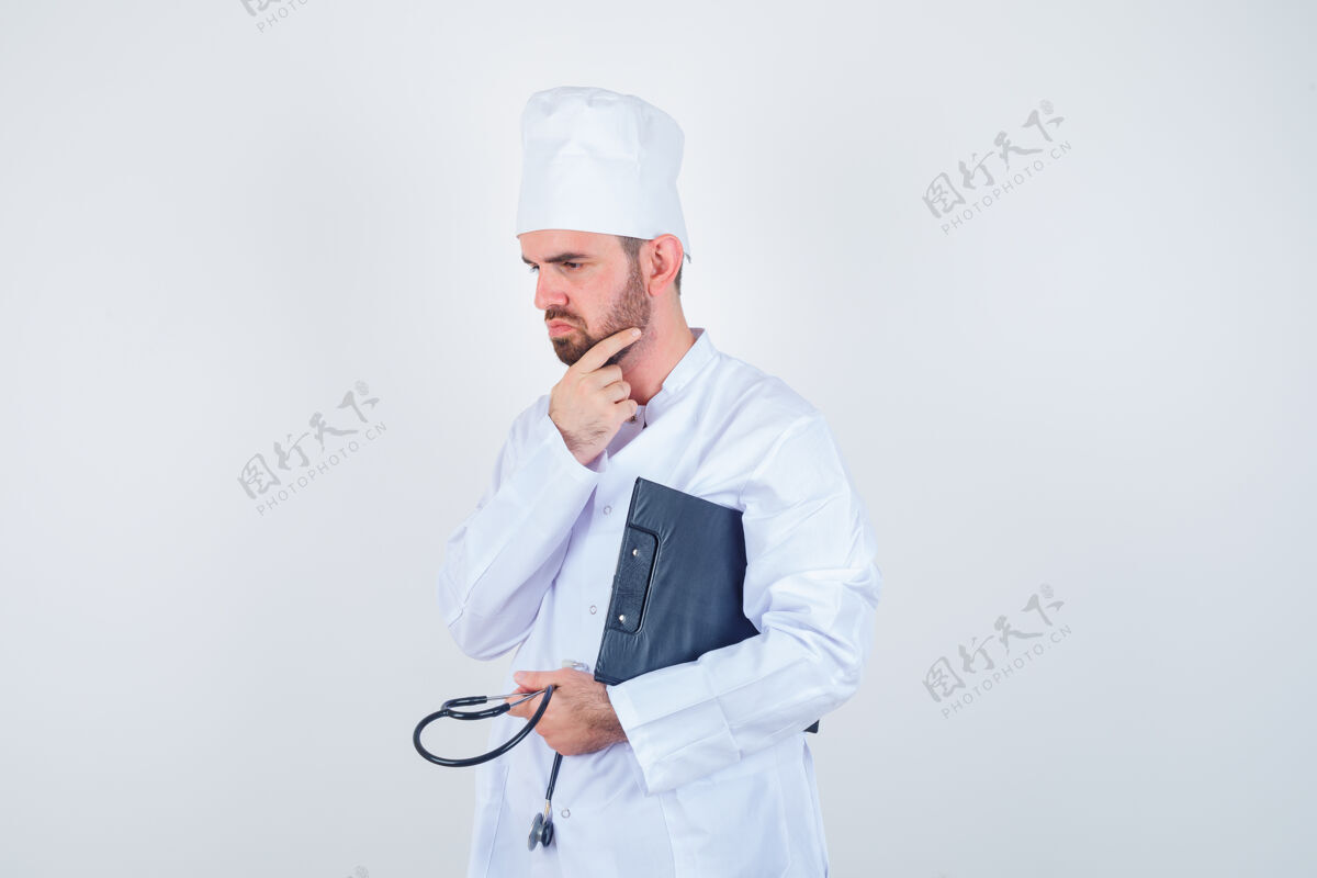 前面年轻男医生拿着剪贴板 听诊器 摸着白制服的胡须 看着若有所思的前视图男人男交叉