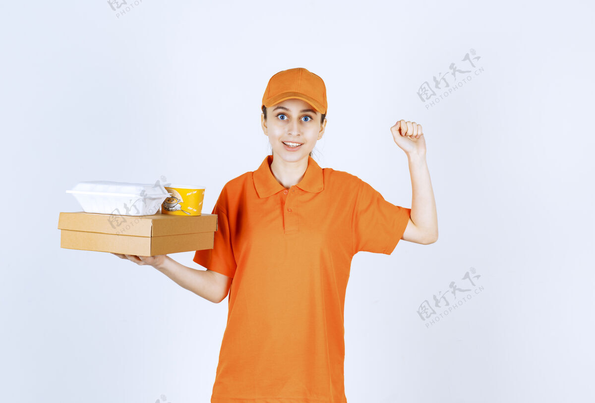 运输身着橙色制服的女快递员手持一个纸板箱 一个塑料外卖箱和一个黄色的面杯 同时显示出积极的手势成功休闲促销