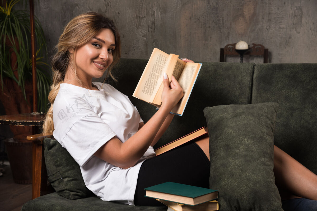 微笑年轻的女人在看一本书 坐在沙发上向前看年轻室内公寓
