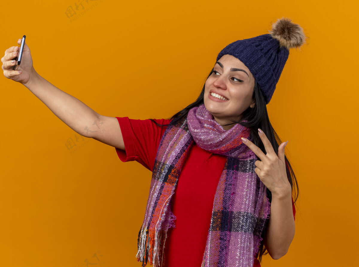 橙色带着微笑的年轻白种人生病的女孩戴着冬天的帽子和围巾自拍做和平标志隔离在橙色的墙上疾病自拍年轻