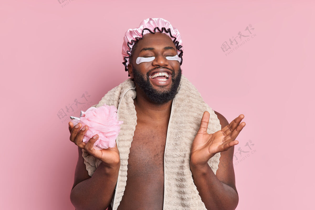 丰满快乐的大胡子男人戴着浴帽在眼睛下面敷上胶原蛋白贴片柔软的毛巾围着脖子拿浴巾享受卫生和美容程序对着粉色的钉墙摆姿势人肖像面部