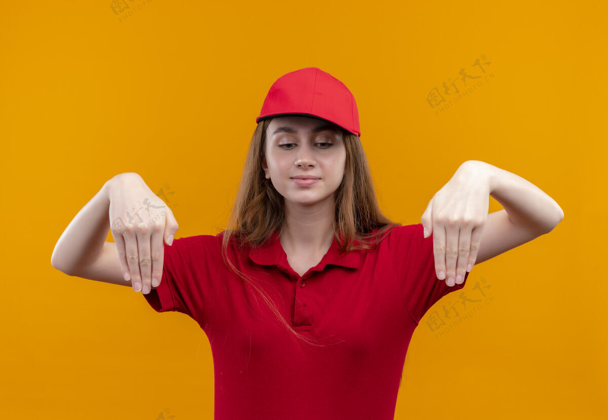手身穿红色制服的年轻送货女 手朝下指着孤立的橙色墙壁年轻女人女孩