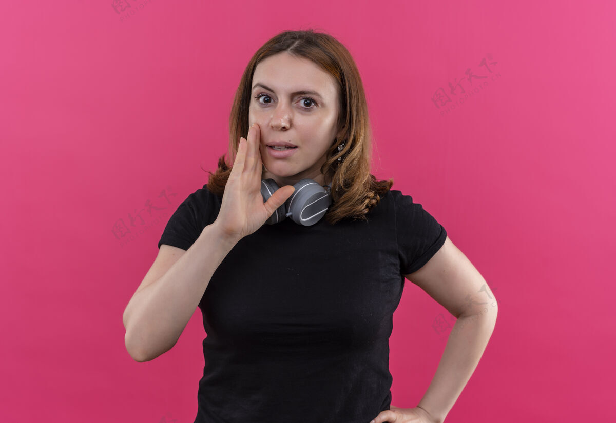 穿在隔离的粉色墙壁上 脖子上戴着耳机 手靠着嘴的年轻休闲女人在低语休闲女人耳机