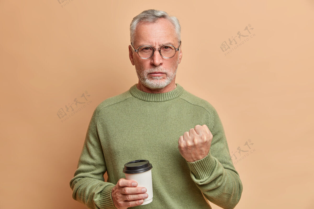 人严厉愤怒的大胡子老人严肃地看着前面试图警告你拿着一次性咖啡杯穿着休闲套头衫对着棕色墙壁摆姿势拳头疯狂欧洲人