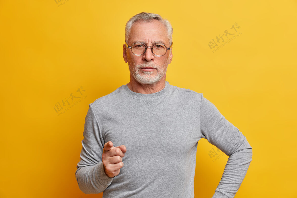 生动严肃严苛的胡须男人画像用手指警告明智的建议穿着灰色长袖套头衫自信地看着前面隔离的黄色墙壁眼镜成熟人