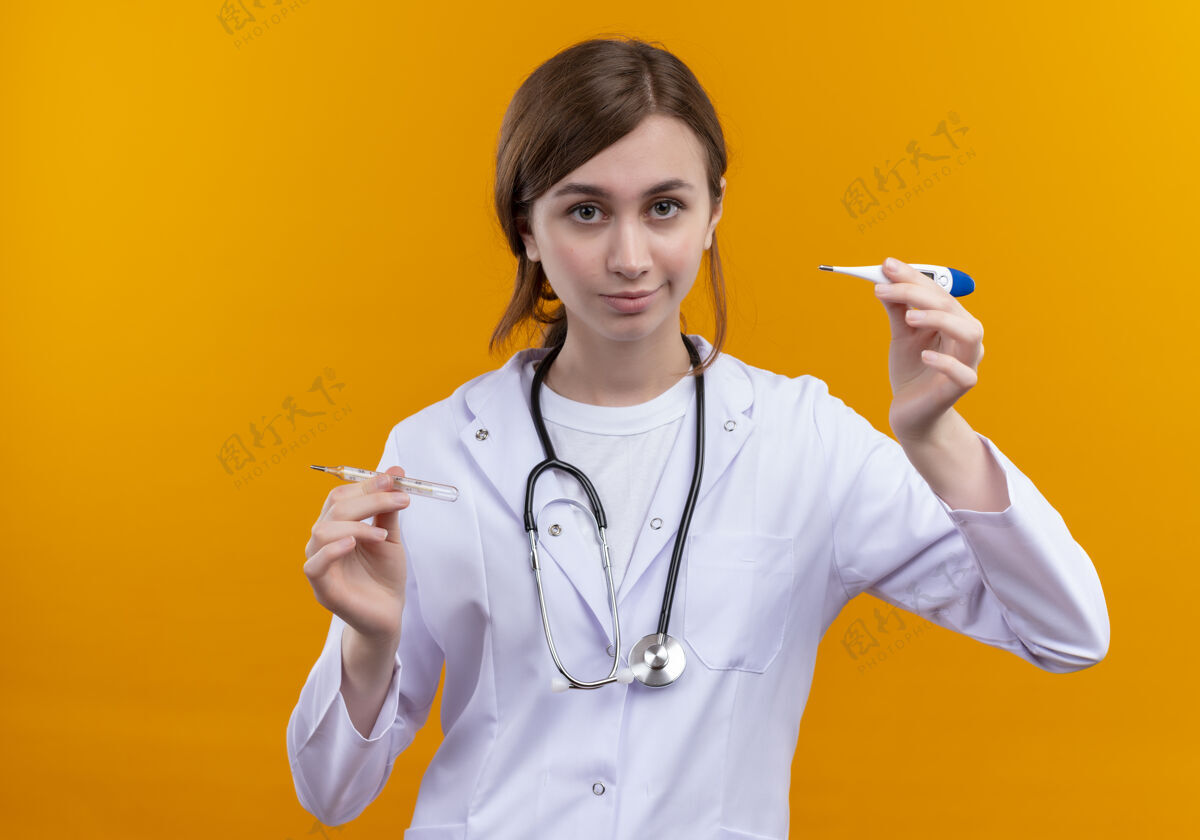 体温计体贴的年轻女医生 穿着医用长袍 戴着听诊器 用手指举着温度计 站在隔离的橙色墙上 留着复印空间女人听诊器拿着