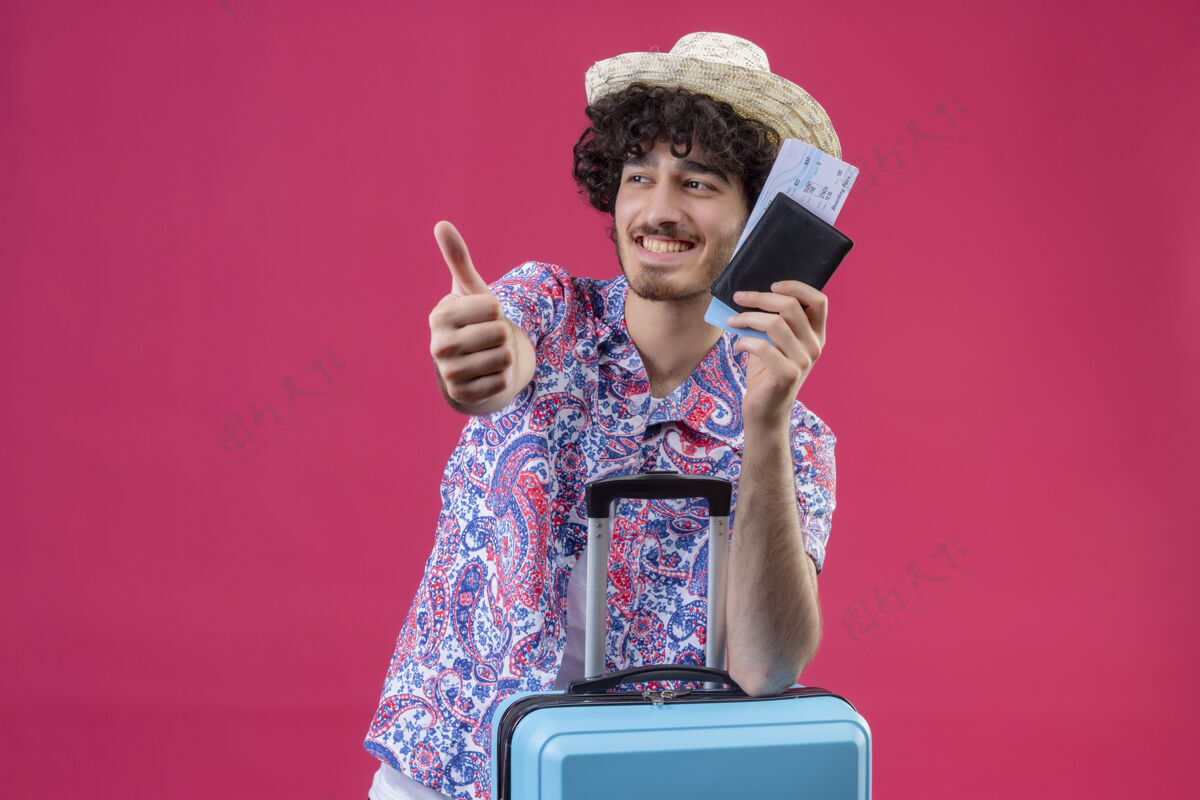 胳膊微笑着的年轻英俊的卷发旅行者戴着帽子拿着钱包和机票 竖起大拇指 把胳膊放在箱子上 放在隔离的粉色墙壁上 有复印空间男人年轻人机票