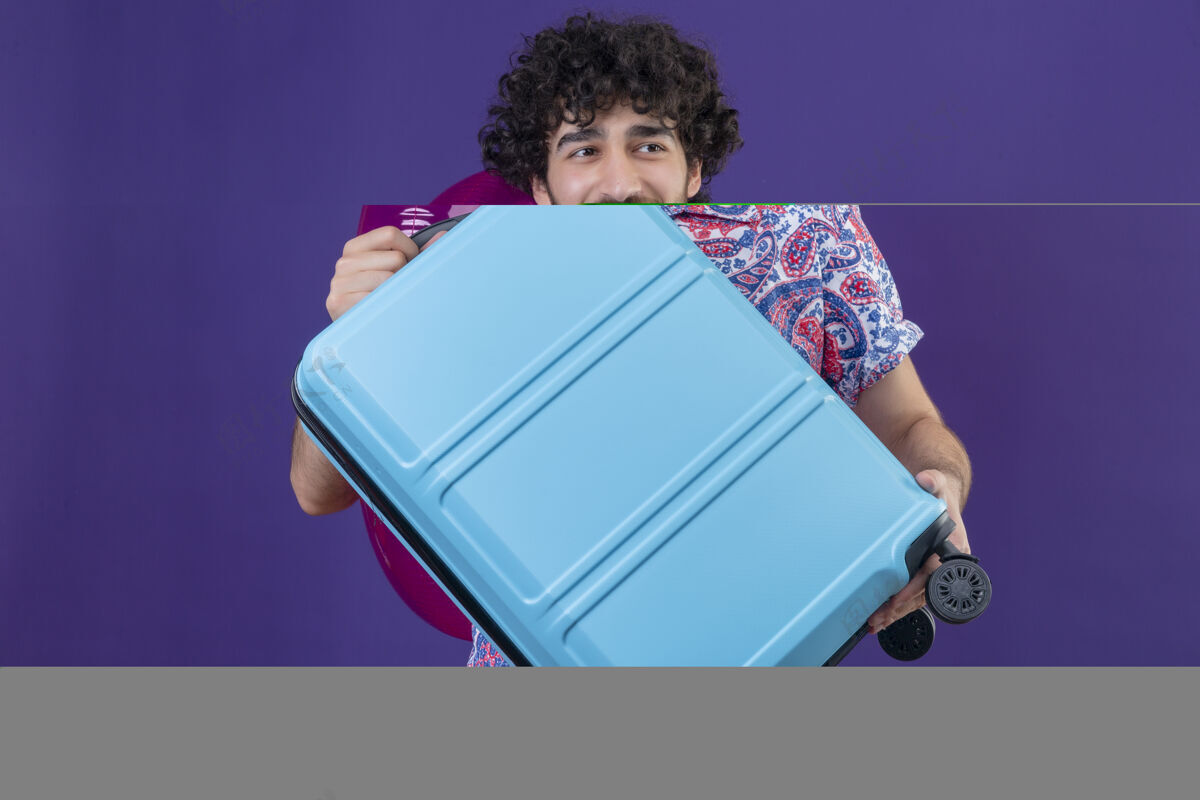 戒指微笑着的年轻英俊的卷发旅行者拿着手提箱和游泳圈在孤立的紫色墙壁上复制空间手提箱男人游泳