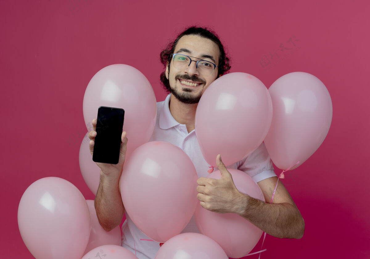 微笑面带微笑的戴眼镜的帅哥站在气球后面 举着电话 他的大拇指孤立地站在粉红色的墙上眼镜电话戴着