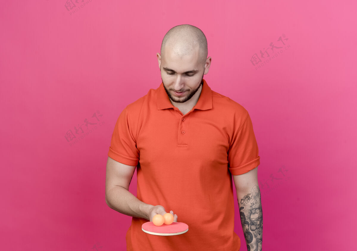 球年轻的运动型男子拿着乒乓球拍 看着球孤立在粉红色的墙上年轻人球拍握