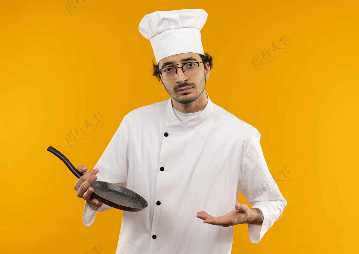 持有年轻的男厨师穿着厨师制服 戴着眼镜 拿着煎锅 把手放在黄色的墙上手眼镜蔓延