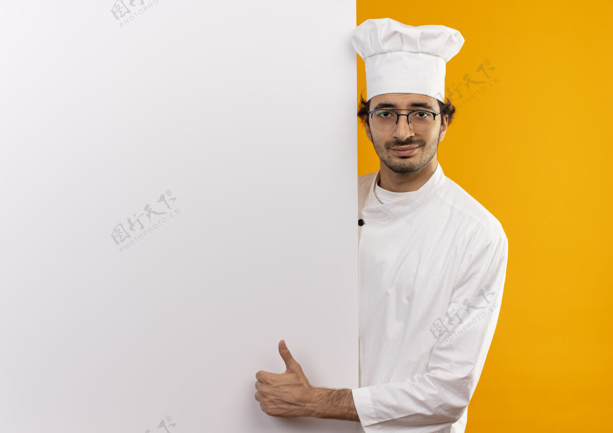 厨师高兴的年轻男厨师穿着厨师制服 戴着眼镜 举着白色的墙壁 他的大拇指孤立地竖立在黄色的墙上 留着复印空间戴着大拇指年轻