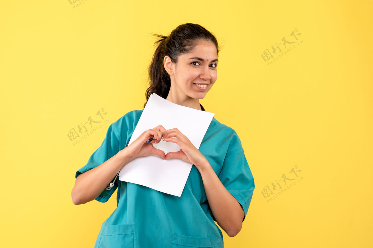 手美丽的女医生的正面图 手放在黄色的墙上 拿着纸做着心的标志微笑肖像人
