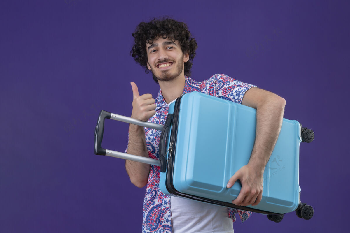 手提箱微笑的年轻英俊的卷发旅行者拿着手提箱 在孤立的紫色墙壁上竖起大拇指 留着复制空间秀帅气卷曲
