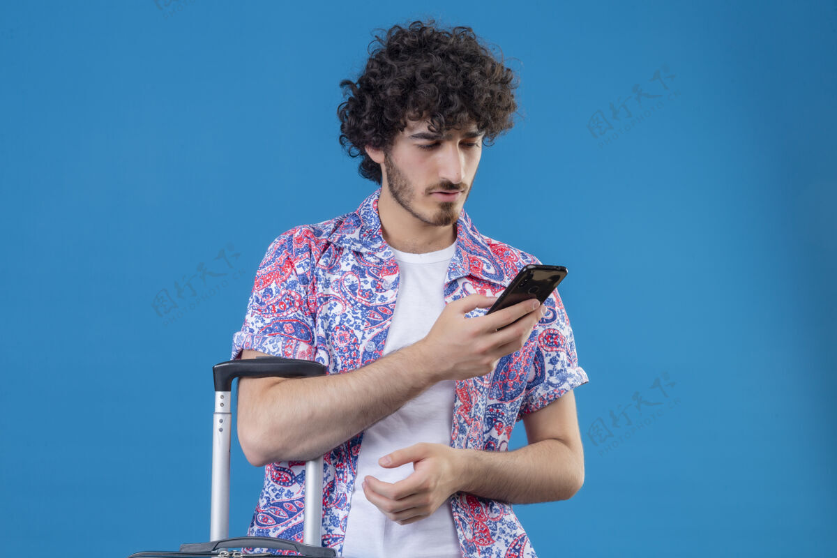 手机年轻英俊的旅行家男子拿着手机 拿着手提箱看着它 在蓝色的隔离墙上留着复印空间帅哥男人电话