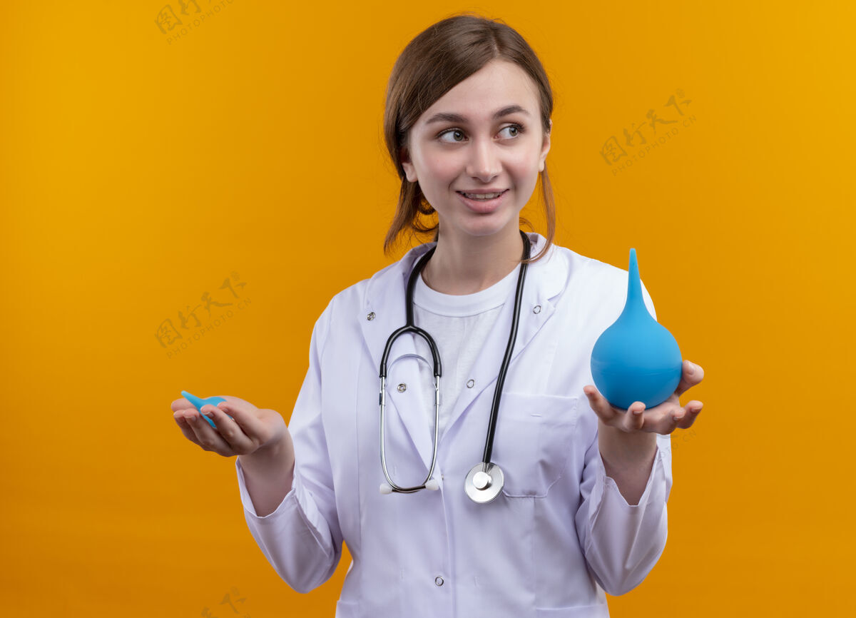 微笑面带微笑的年轻女医生穿着医用长袍 手持听诊器在隔离的橙色墙上灌肠穿女人医生