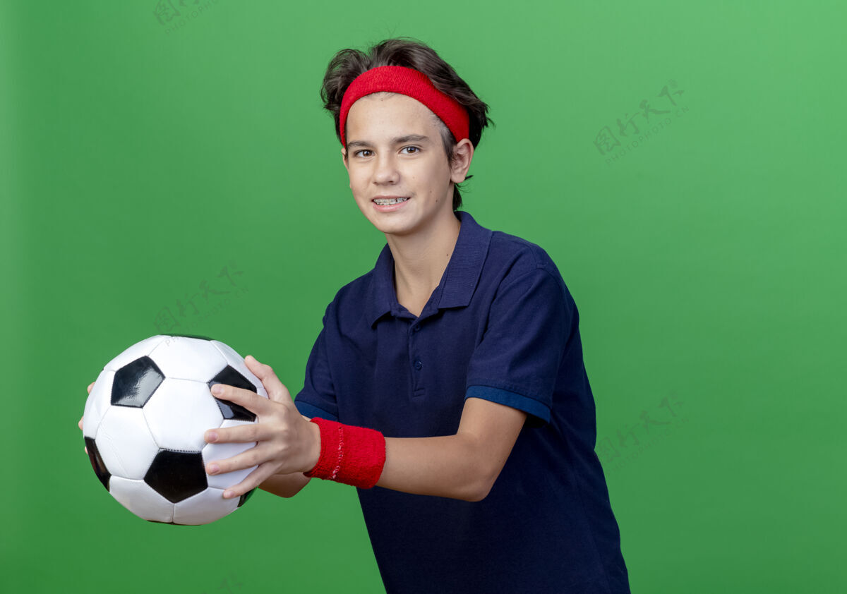 年轻面带微笑的年轻帅气的运动男孩戴着头带和护腕 戴着牙套 足球被隔离在绿色的墙上 留有复制空间牙齿帅气头带