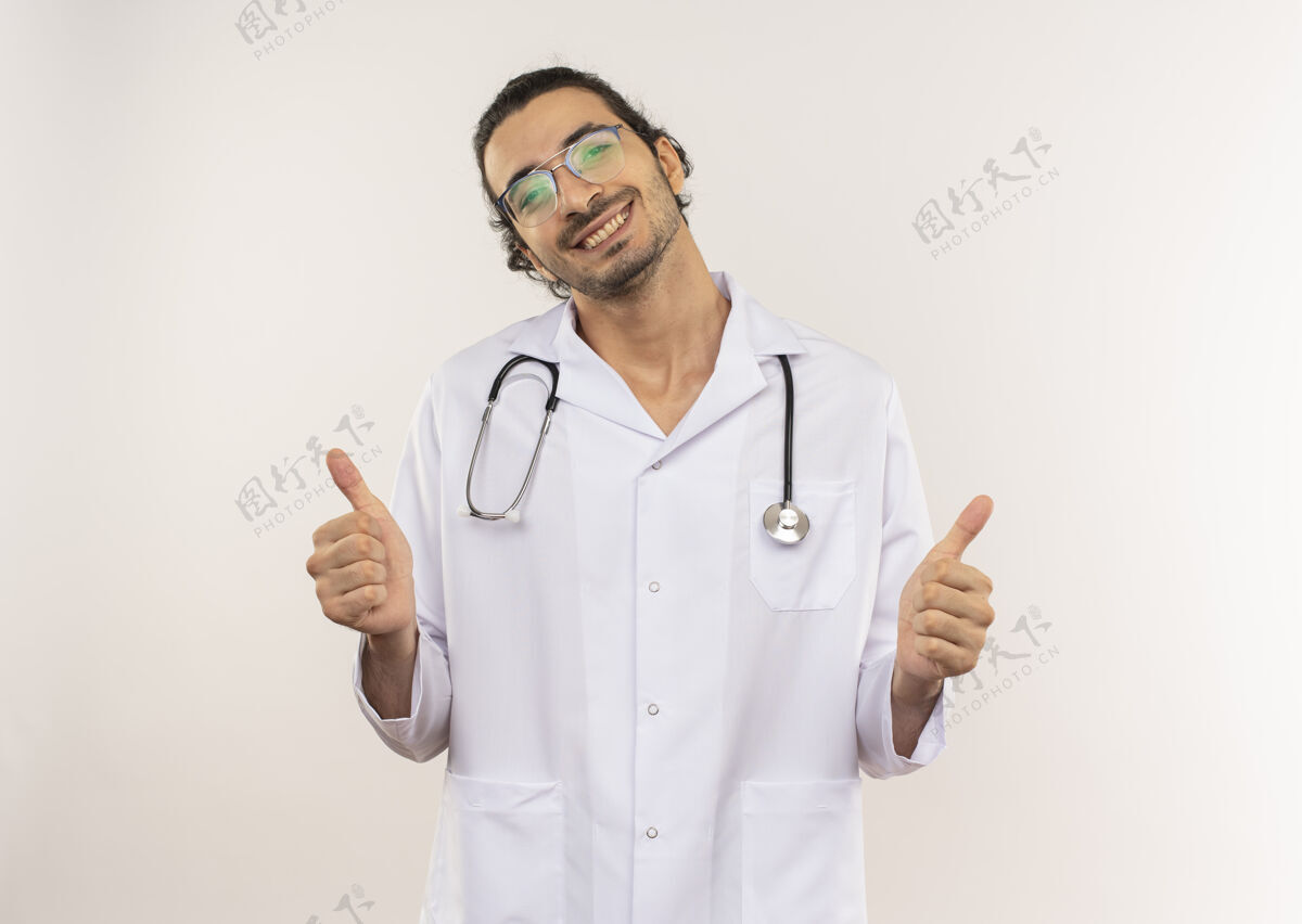 光学微笑的年轻男医生戴着眼镜 穿着白色长袍 带着听诊器 他竖起大拇指站在隔离的白色墙壁上 留着复印空间长袍拇指微笑