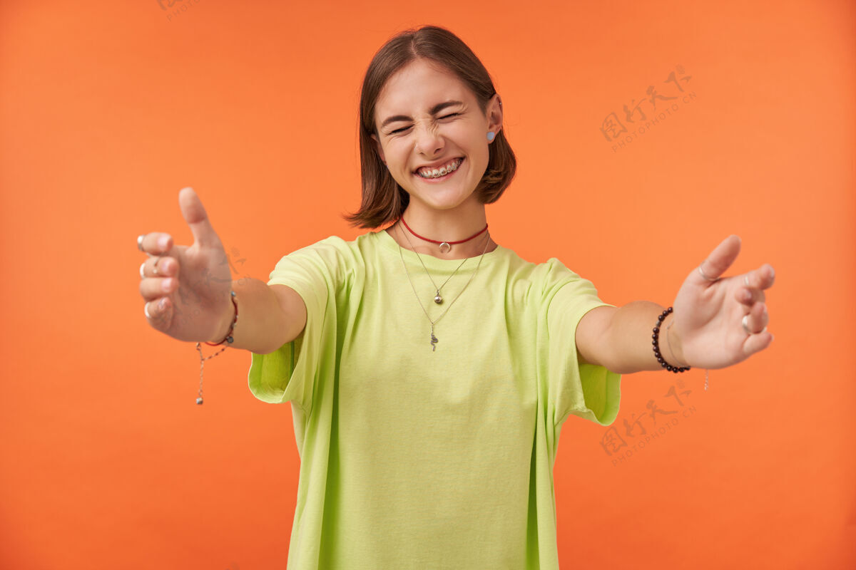 微笑年轻漂亮的女人眯着眼睛微笑 伸出手来拥抱学生很高兴看到她的朋友们穿着绿色t恤 戴着牙套 手镯 项链橙色墙上的肖像T恤休闲女性