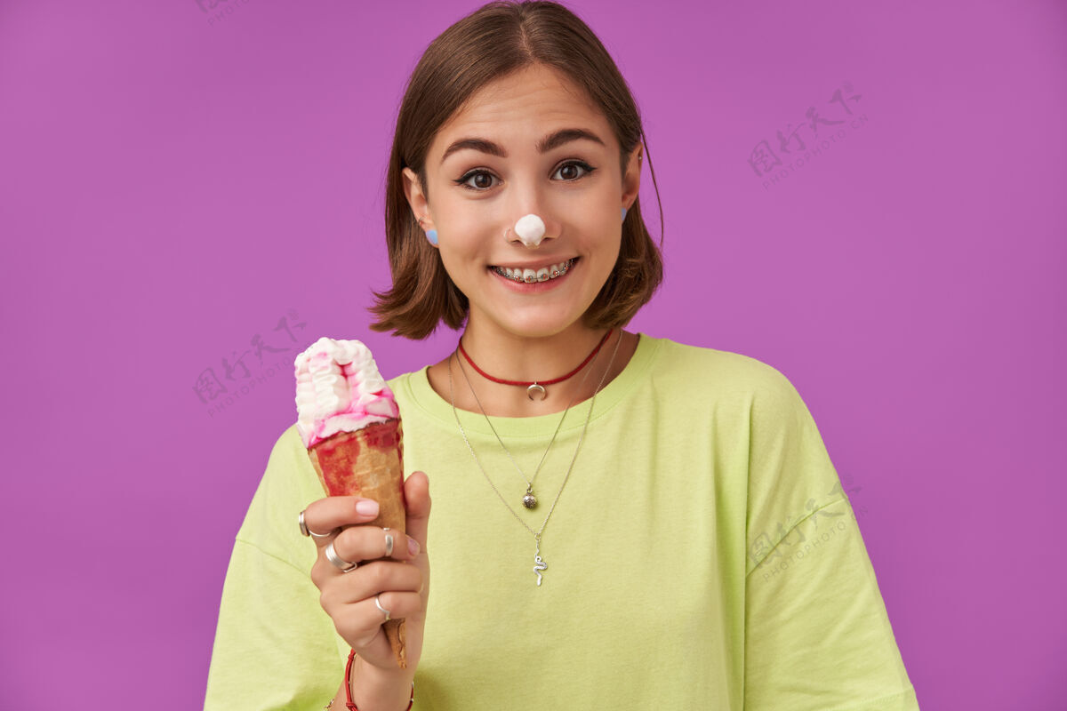 成人十几岁的女孩 快乐快乐 深色短发 鼻子上放着冰激凌拿着冰激凌微笑着 越过紫色的墙穿着绿色t恤 戴着牙套和手镯穿洞牙齿20多岁