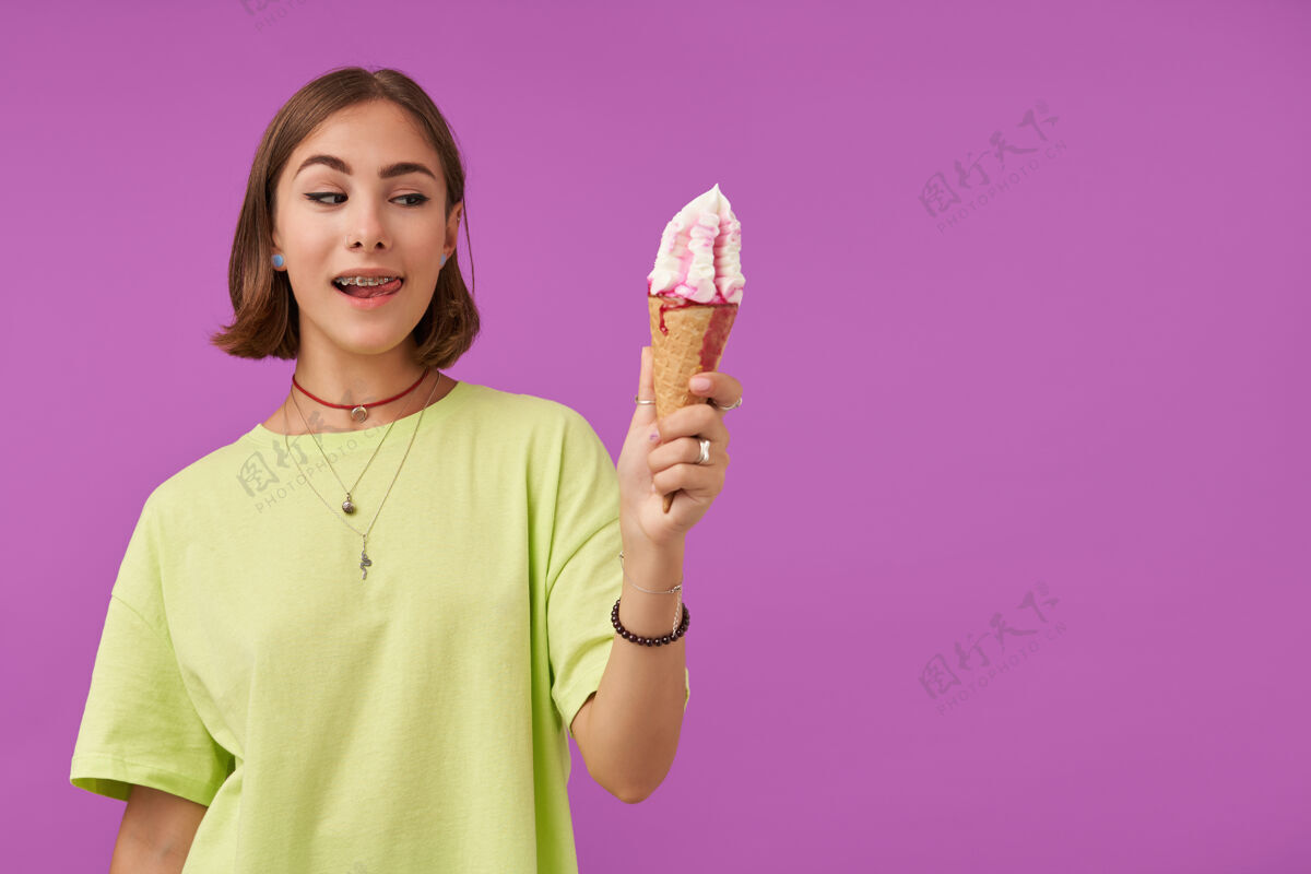 女性漂亮漂亮的女孩的肖像看着冰淇淋 露出舌头在紫色墙上的右边空白处复制穿着绿色t恤 手镯 戒指和项链圆锥形欢乐冰淇淋