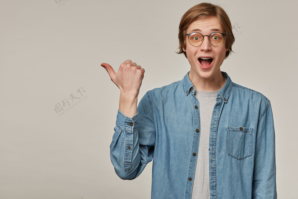 背带惊讶的家伙 快乐的金发男人穿着蓝色牛仔衬衫 戴着眼镜 戴着牙套拇指指向左边的复印空间 隔着灰色的墙颜色室内眼镜