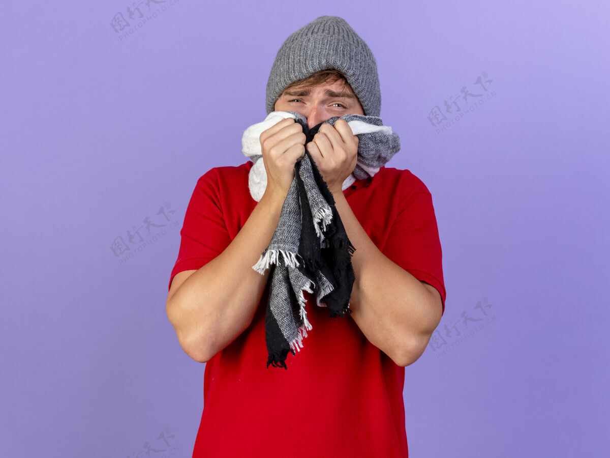 英俊弱小的年轻英俊的金发病夫戴着冬天的帽子和围巾看着相机用围巾遮住嘴看着相机隔离在紫色背景上的复制空间掩护相机围巾